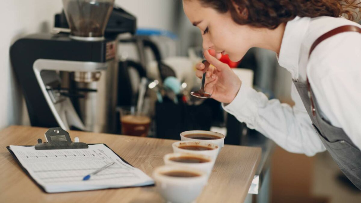 A woman tasting coffee from a spoon with cups and a clipboard on the table.